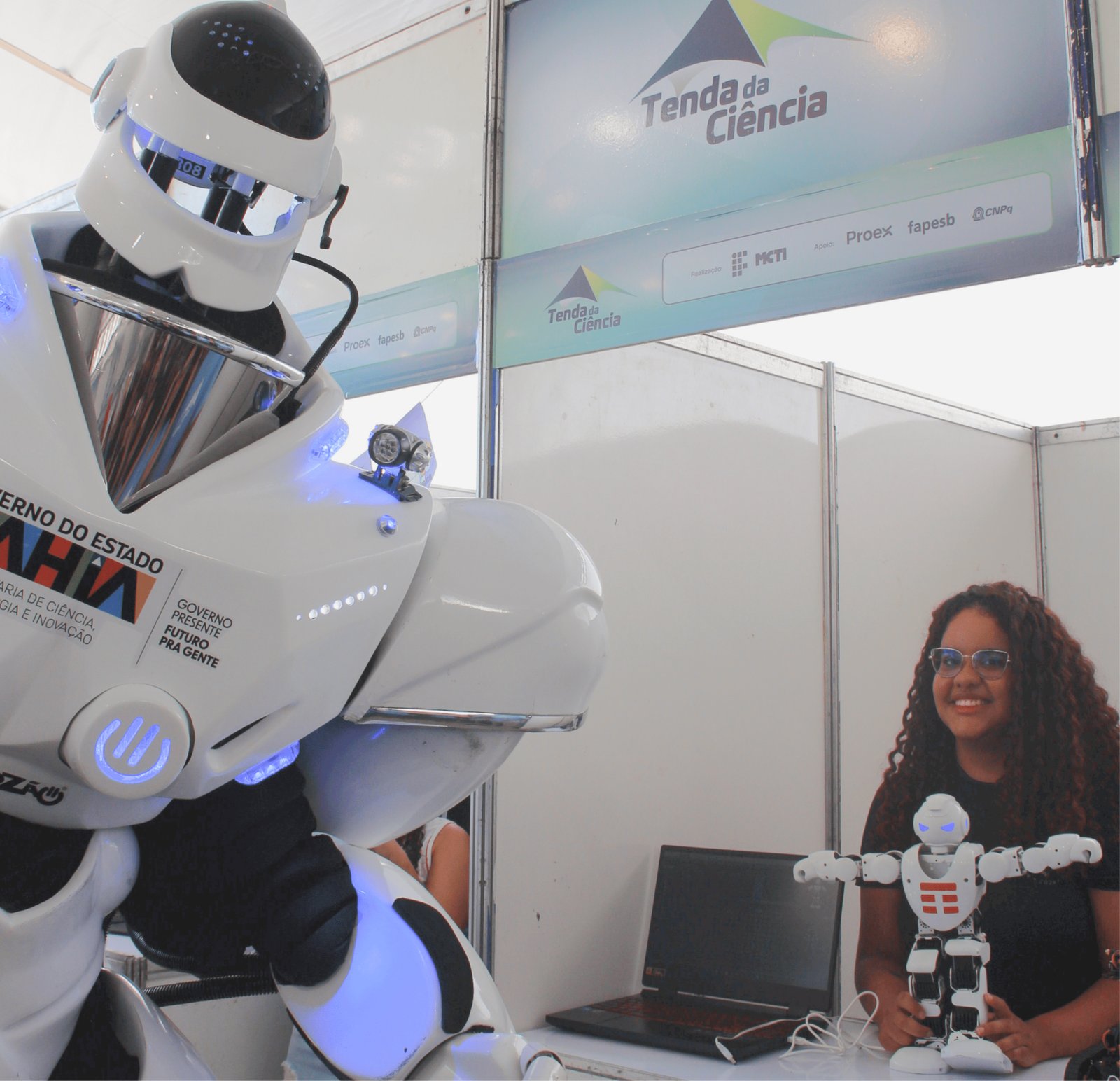 Uma jovem de óculos e longos cabelos cacheados sorri sentada em um estande. Ao seu lado esquerdo, está o enorme Robozão branco (em escala humana). À sua frente, sobre a mesa, ela segura um pequeno robô humanoide branco e preto (escala de mesa) ao lado de um notebook preto. O contraste entre os dois robôs destaca as diferentes aplicações da robótica apresentadas no evento.