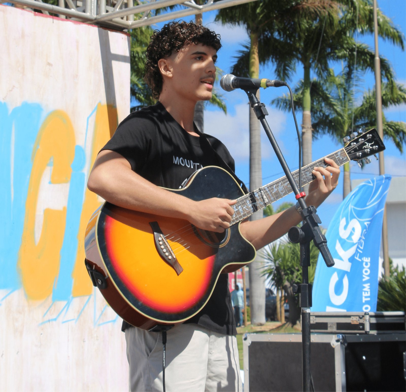 Um rapaz de cabelos cacheados e camiseta preta toca um violão eletroacústico sunburst (degradê de amarelo para preto). Ele canta em um microfone montado em um pedestal. O cenário é externo, com palmeiras altas ao fundo, um céu azul límpido e uma bandeira azul promocional com o texto "HICKS FIBRA". Ele parece estar em uma apresentação solo no palco da feira.