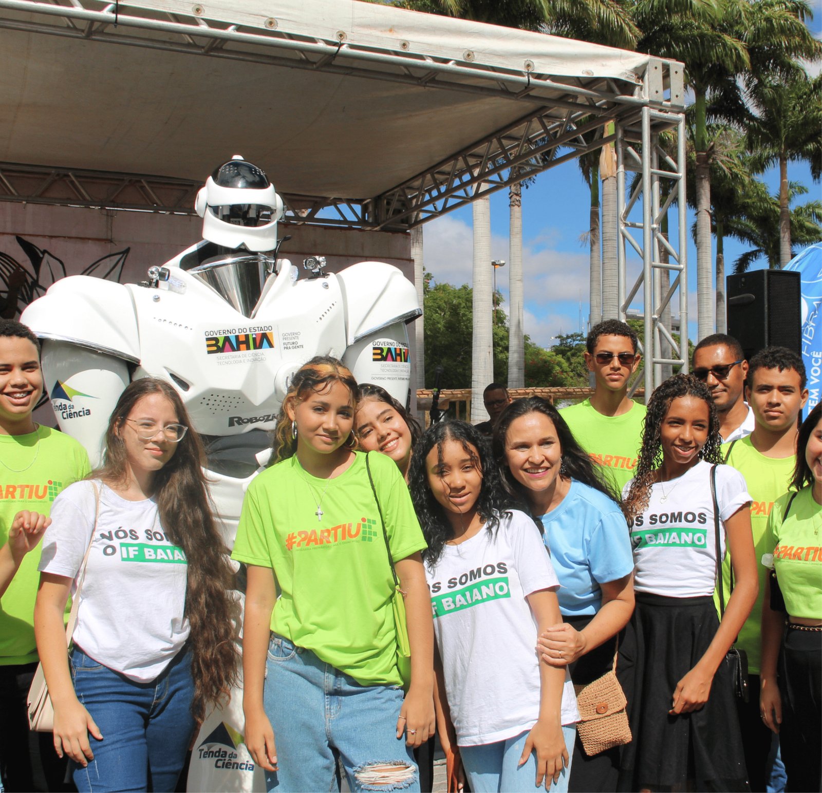 Um grupo de 10 estudantes e uma professora posam para uma foto oficial com o grande Robozão branco ao fundo. Metade do grupo usa camisetas verde-limão vibrante com a hashtag "#PARTIU", e a outra metade usa camisetas brancas com a frase "NÓS SOMOS IF BAIANO". Eles estão em um palco e o clima é de união e orgulho institucional. Palmeiras e o céu azul completam o cenário de fundo.