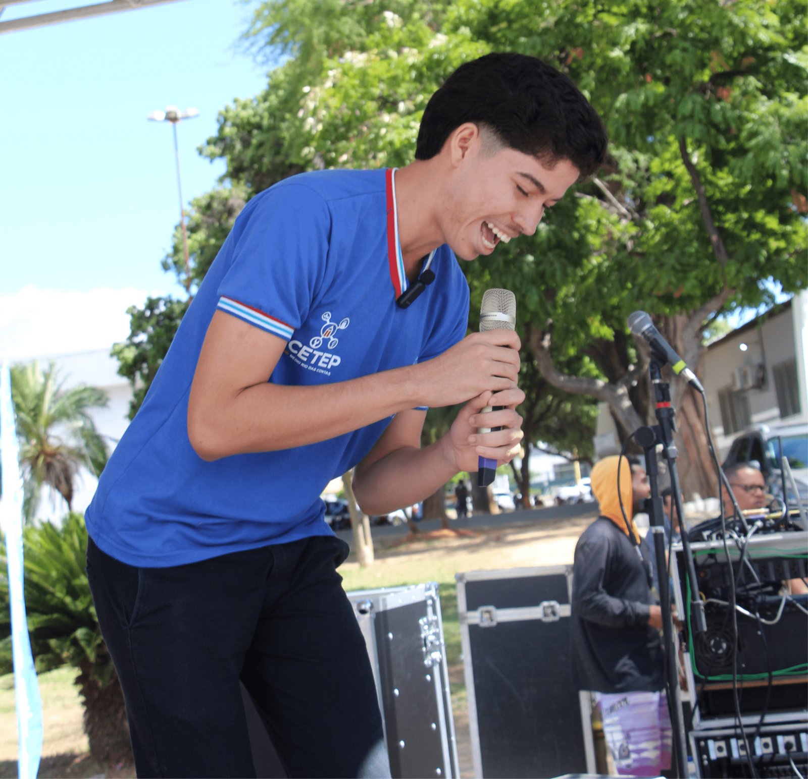 Um jovem de perfil, vestindo uma camisa polo azul do "CETEP", segura um microfone com as duas mãos e ri abertamente, parecendo estar contando uma piada ou celebrando um momento no palco. Ele tem um corte de cabelo moderno, curto nas laterais. Ao fundo, árvores verdes e equipamentos de som pretos indicam a estrutura de um evento festivo ao ar livre.