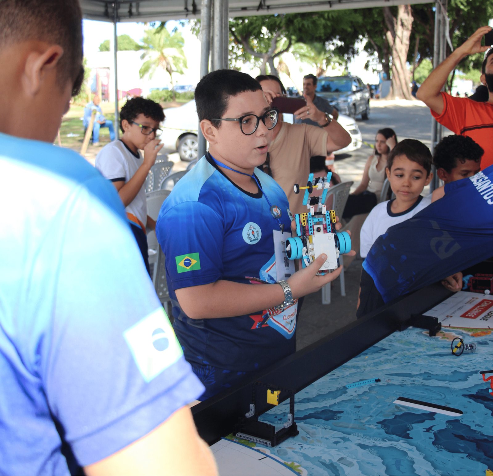 Close-up de um menino de pele clara, cabelos pretos curtos e óculos de armação preta grossa. Ele veste uma camisa técnica azul com detalhes em degradê e a bandeira do Brasil na manga. Com as duas mãos, ele segura cuidadosamente um robô construído com peças de LEGO (azul, amarelo e branco) com rodas grandes. Ele parece estar explicando o projeto, com a boca entreaberta. Ao fundo, o ambiente de feira está movimentado com pessoas tirando fotos.