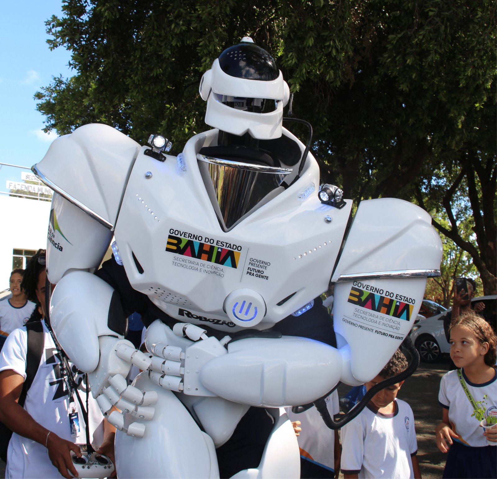 Imagem em ângulo baixo destacando um robô humanoide de aproximadamente 2 metros de altura, feito de material plástico branco brilhante com detalhes em preto e luzes LED azuis. No peito, ostenta a marca colorida "GOVERNO DO ESTADO BAHIA" e "Secretaria de Ciência, Tecnologia e Inovação". Pequenas luzes de LED brilham nos ombros. Crianças ao redor olham para cima com admiração; uma menina à direita segura uma pequena muda de planta em um copinho plástico.