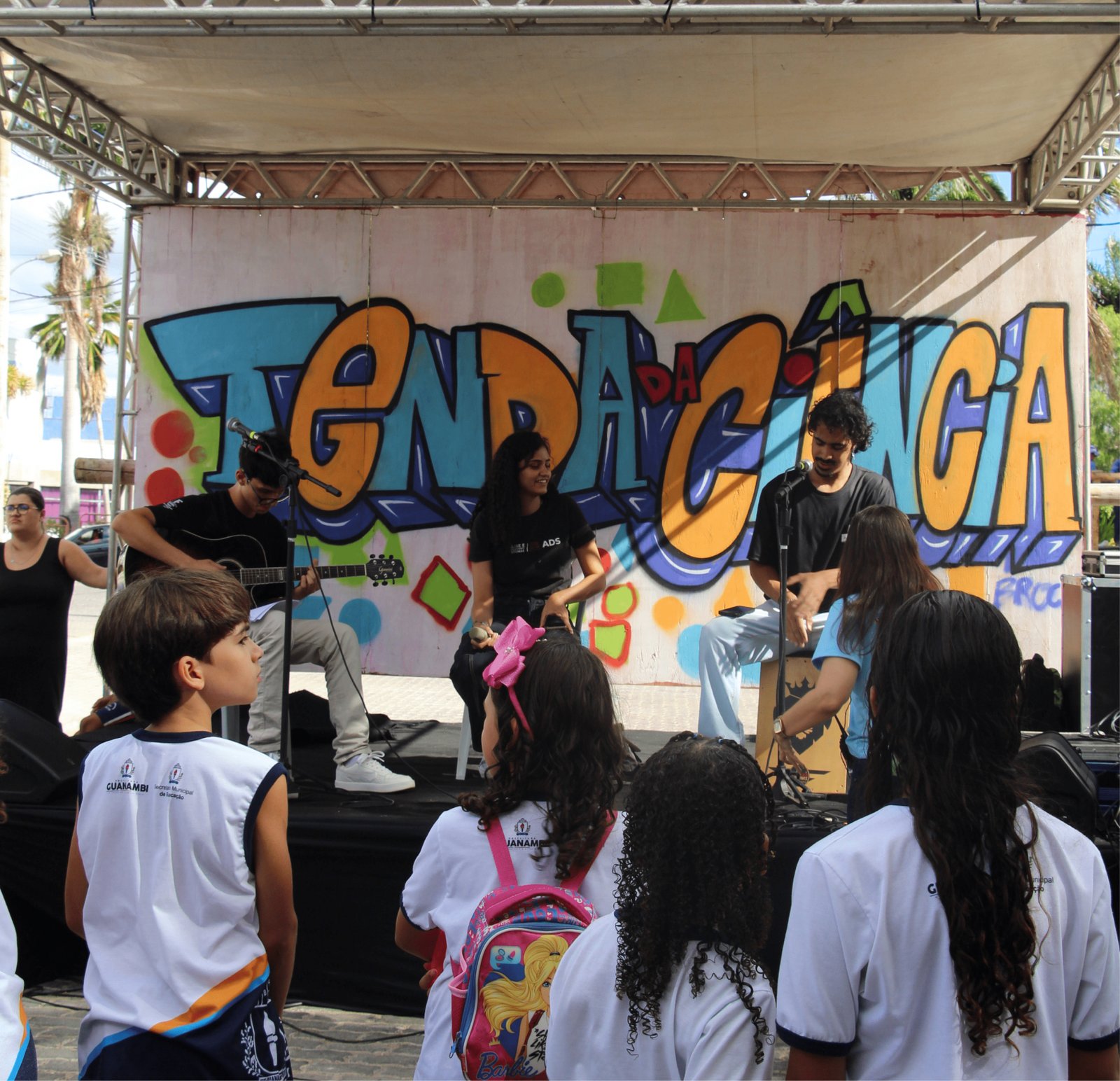 Vista de um palco baixo sob uma tenda. Ao fundo, um painel de madeira exibe um grafite artístico com letras grandes e coloridas (azul, laranja e amarelo) onde se lê "TENDA DA CIÊNCIA". Três jovens se apresentam: um rapaz à esquerda toca violão preto, uma moça ao centro canta com um microfone e um rapaz à direita toca um cajón. Em primeiro plano, vemos as costas de crianças com uniformes escolares brancos da rede municipal de Guanambi observando o show.