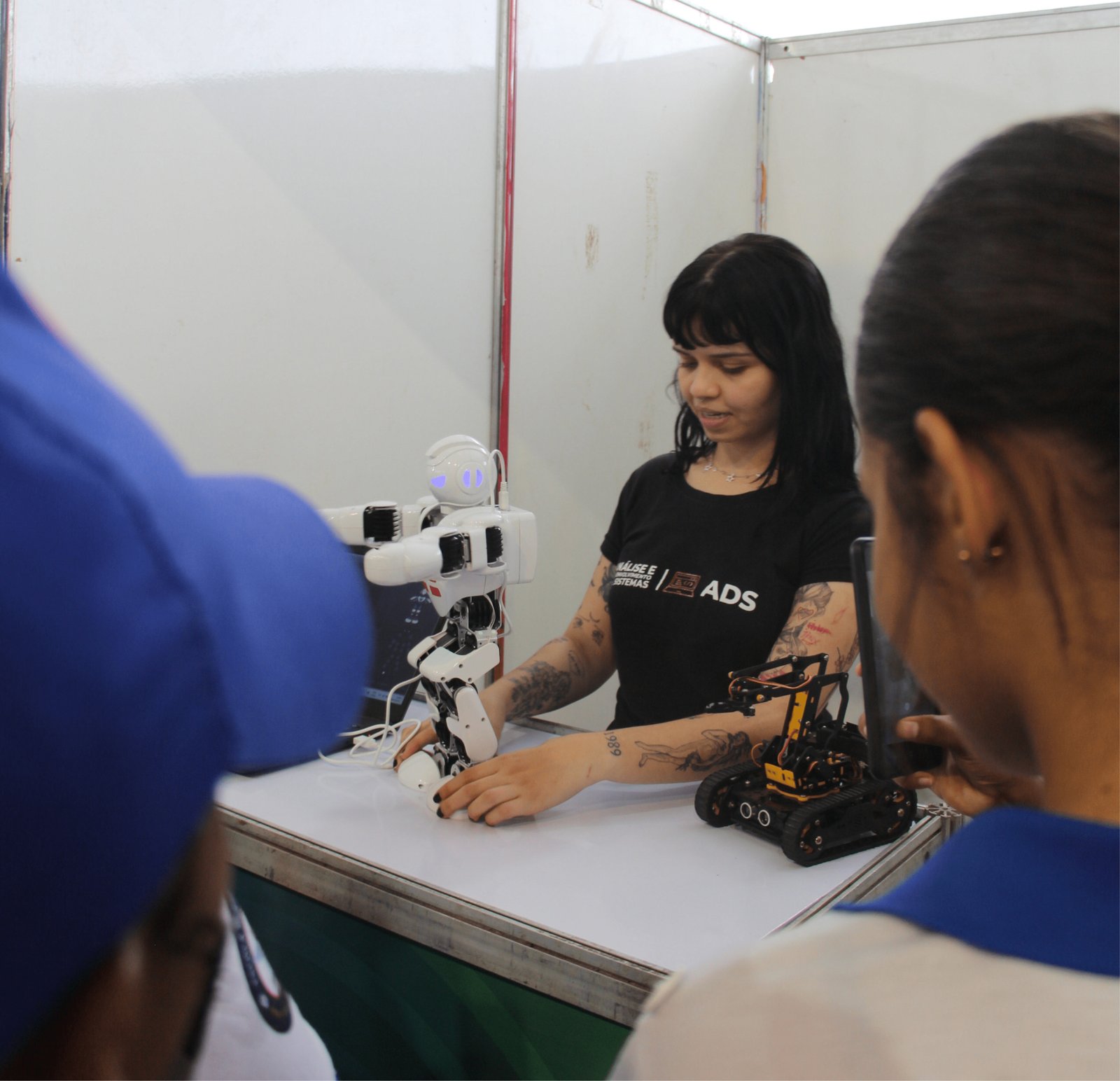 Uma jovem de cabelos pretos e braços cobertos por tatuagens artísticas está atrás de um balcão branco. Ela veste uma camiseta preta com a sigla "ADS" (Análise e Desenvolvimento de Sistemas). À sua frente, há um robô humanoide branco pequeno em posição de saudação e um pequeno robô de esteira preto e amarelo. Visitantes em primeiro plano (desfocados) observam a demonstração. O ambiente é um estande de feira com divisórias brancas.