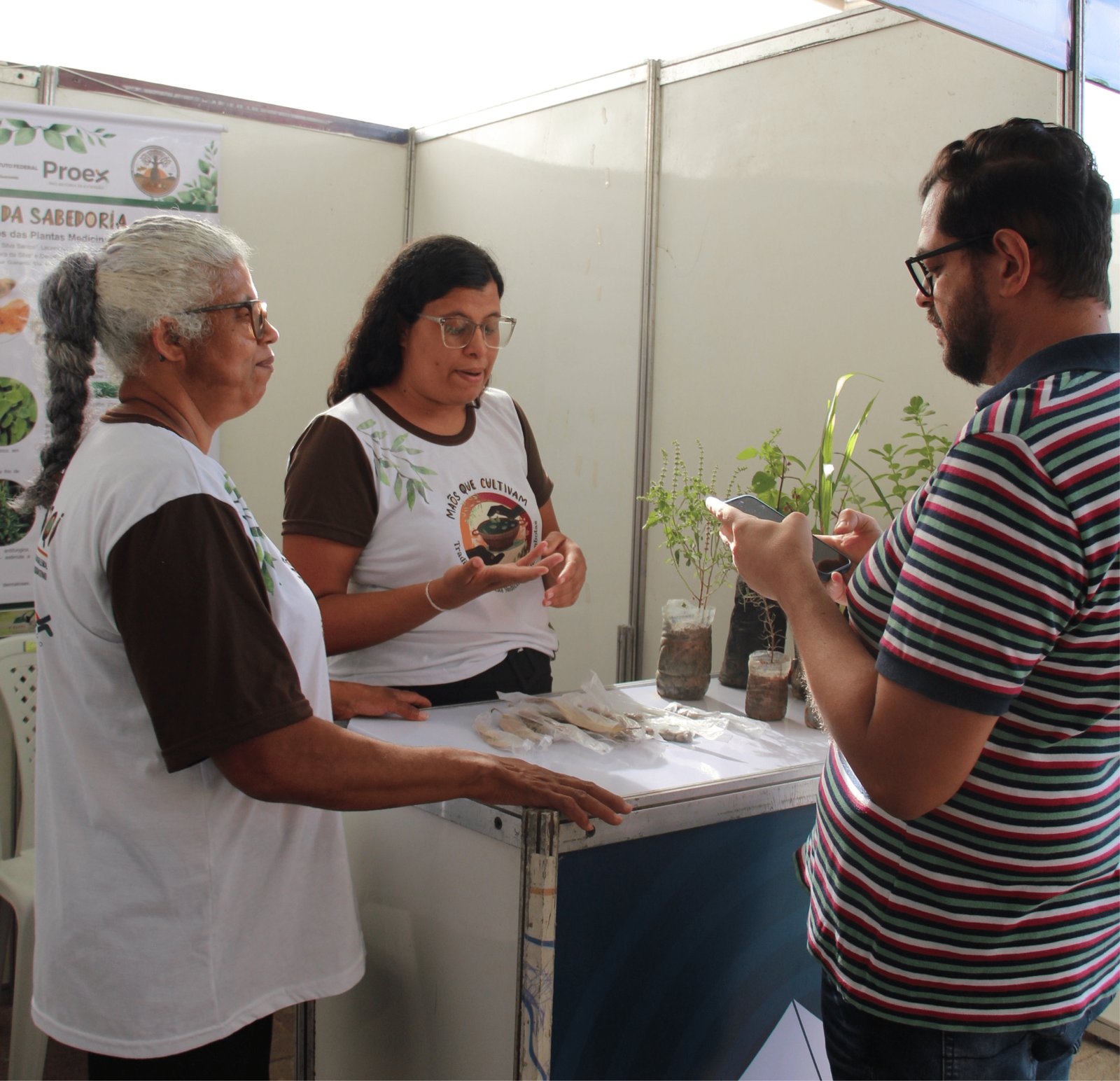 Em um estande, uma senhora negra com longas tranças grisalhas presas e óculos conversa com um homem de camisa listrada. Ela veste uma camiseta branca com o logotipo "Sabedoria das Plantas Medicinais". Ao lado dela, uma jovem de óculos gesticula explicando o projeto. Sobre o balcão, há pequenos vasos com mudas verdes e amostras de plantas desidratadas em saquinhos plásticos. O homem usa o celular para fotografar os detalhes.