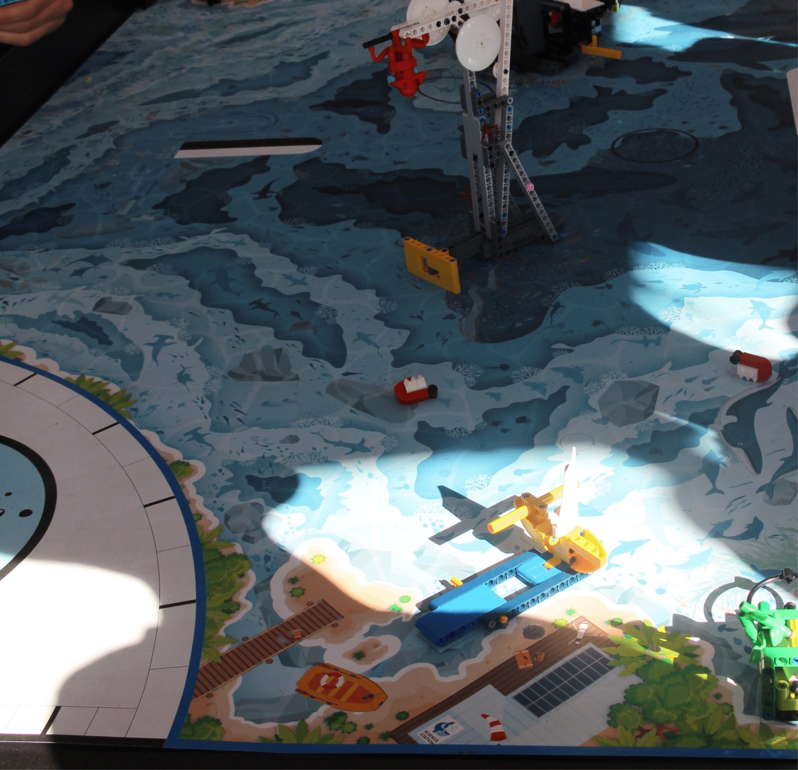 Vista detalhada de um tapete plástico de competição de robótica. O design imita o fundo do mar e uma costa litorânea em tons de azul e areia. Sobre o tapete, há estruturas construídas com LEGO: uma turbina eólica cinza, um pequeno barco vermelho e branco, uma plataforma amarela e um veículo subaquático amarelo e azul. O tapete está cheio de linhas e marcas geométricas para orientação de robôs autônomos.