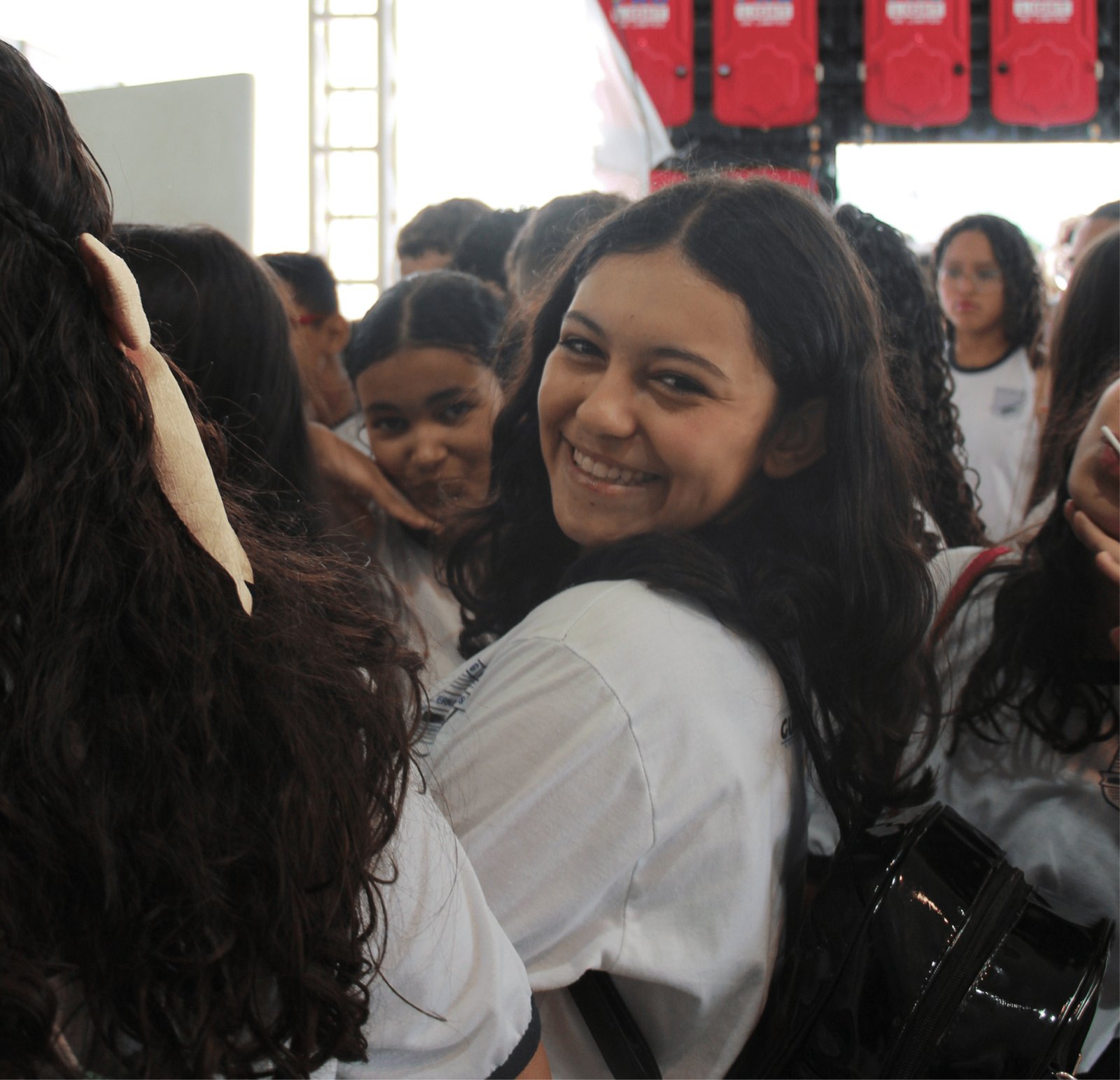 Um close-up focado no rosto de uma adolescente negra que sorri lateralmente para a câmera. Ela tem cabelos longos e cacheados e veste uma camiseta branca de uniforme. A profundidade de campo é rasa, deixando o fundo (com outros estudantes e estruturas vermelhas) desfocado. Ela transmite uma expressão de felicidade genuína e espontaneidade.