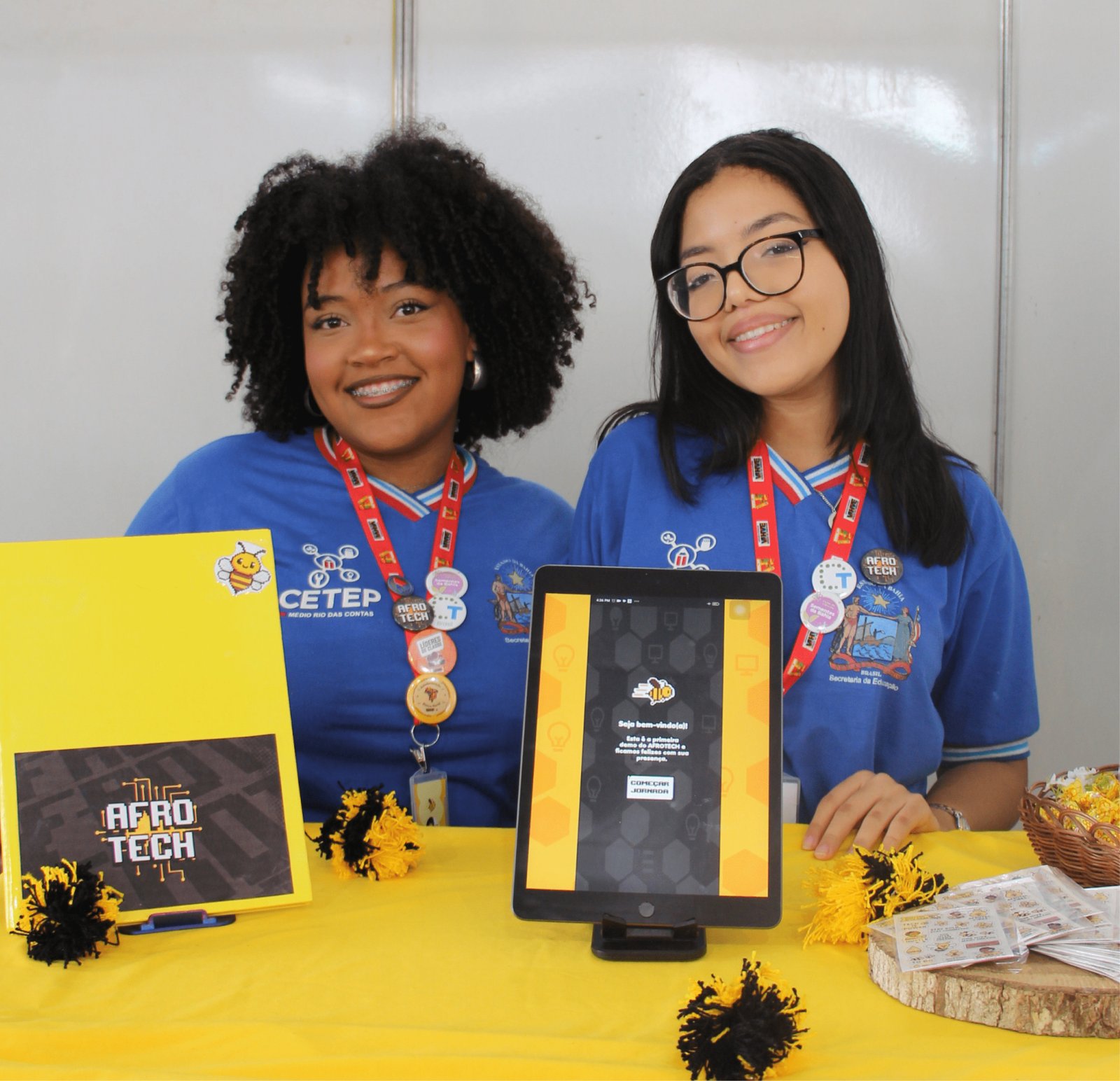 Duas jovens negras sorridentes estão atrás de uma mesa coberta com tecido amarelo vibrante. A da esquerda tem cabelo afro volumoso e usa aparelho; a da direita usa óculos e cabelos lisos. Ambas vestem camisas polo azuis do "CETEP Médio Rio das Contas". Elas apresentam o projeto "AFRO TECH", simbolizado por uma abelha robótica amarela. Um tablet no centro exibe a tela inicial de um aplicativo com a mensagem "Seja bem-vindo(a)".