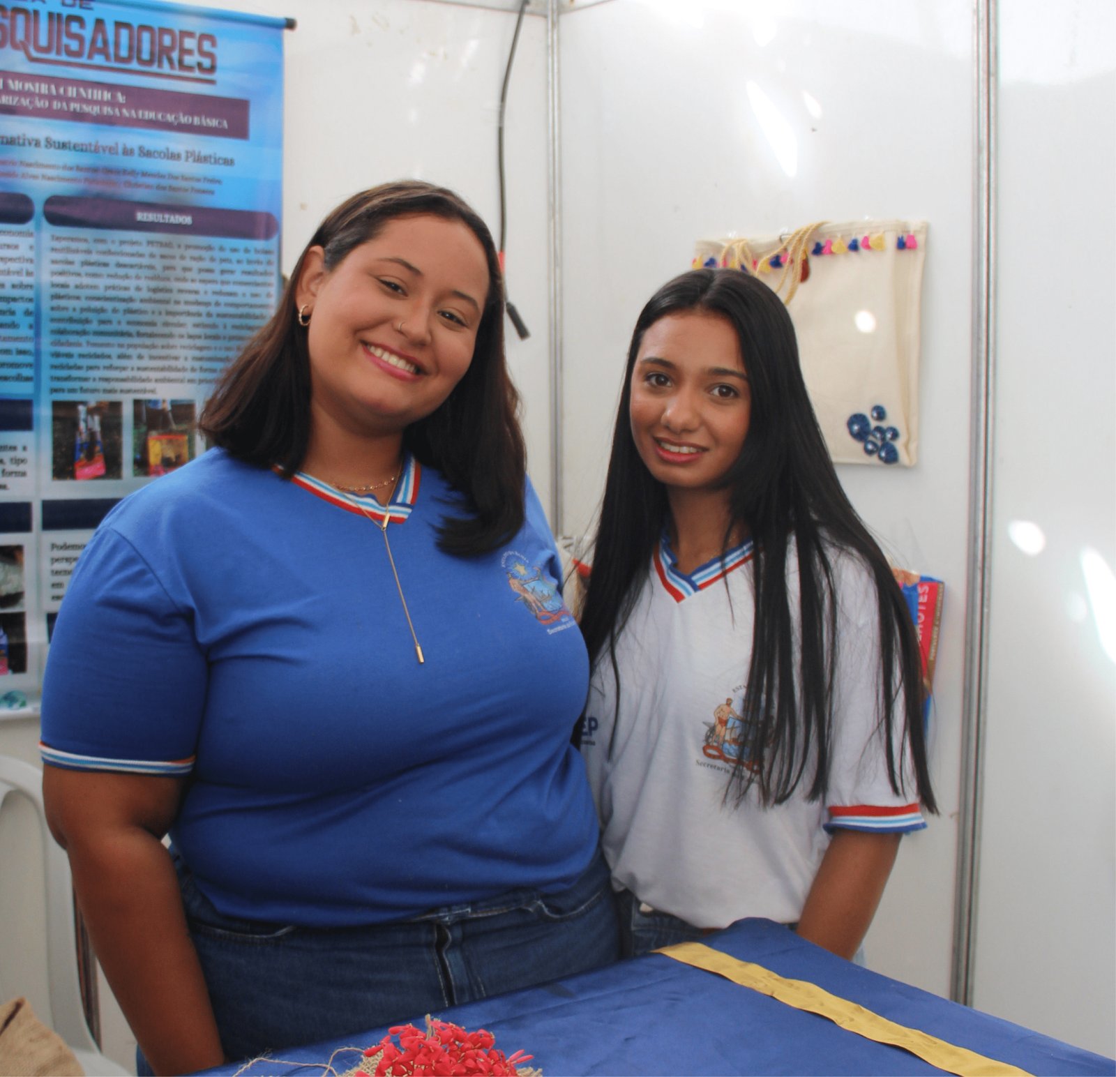 Duas estudantes sorriem ao lado de um banner de pesquisa. A jovem à esquerda é mais robusta, tem cabelos escuros ondulados e usa uma camiseta azul com detalhes em vermelho e branco na gola. A jovem à direita é mais magra, tem cabelos pretos longos e lisos e veste uma camiseta branca. O banner atrás delas contém textos técnicos e fotos de áreas verdes, com o título "Alternativa Sustentável às Sacolas Plásticas".