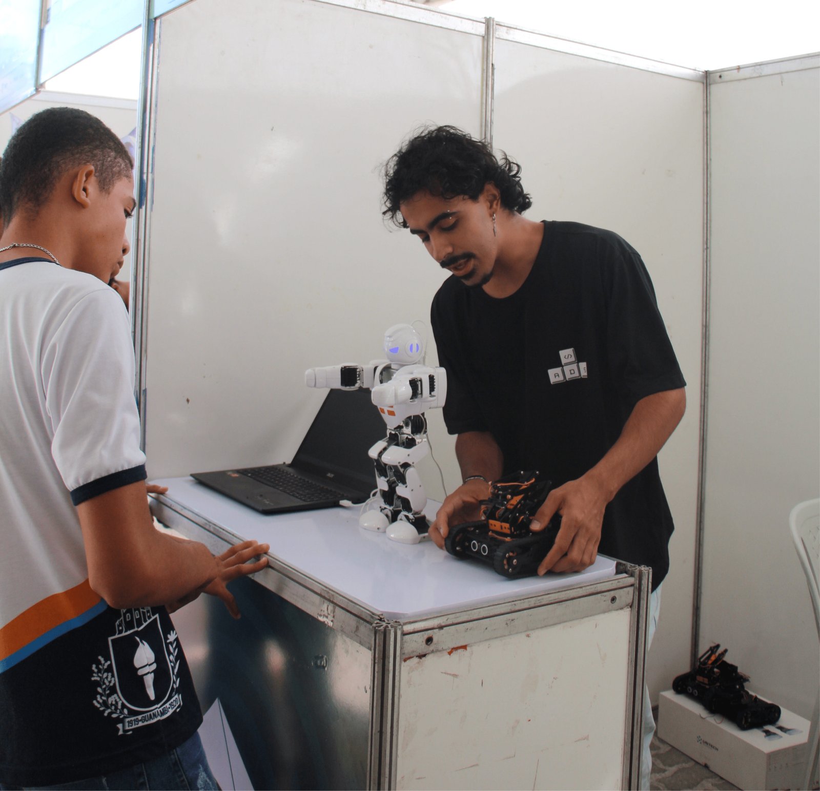 Um jovem de camiseta preta com um logotipo de teclas de computador "A D S" inclina-se sobre um balcão para ajustar um pequeno robô de esteira preto. Ele tem cabelos cacheados e usa um brinco pequeno. À esquerda, um estudante de uniforme escolar observa atentamente. No balcão, há também um robô humanoide branco e um notebook preto aberto.
