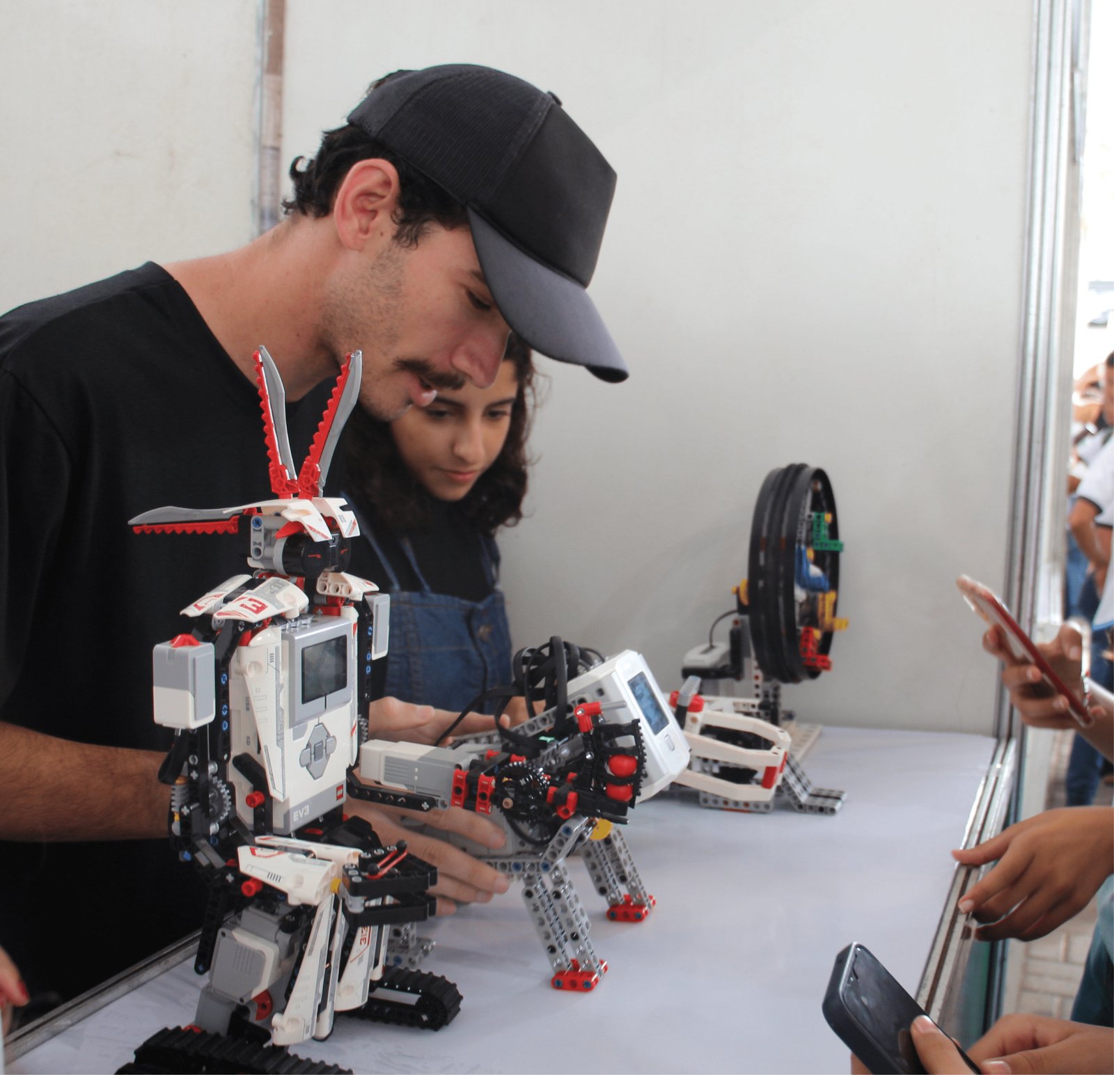 Um rapaz de boné preto e camiseta preta observa atentamente um robô feito de LEGO que lembra um coelho, com "orelhas" vermelhas longas. Ao lado dele, uma menina também observa com foco. Na mesa branca, há outros mecanismos de robótica educativa. Ao fundo, mãos de outros visitantes seguram celulares, registrando a tecnologia.