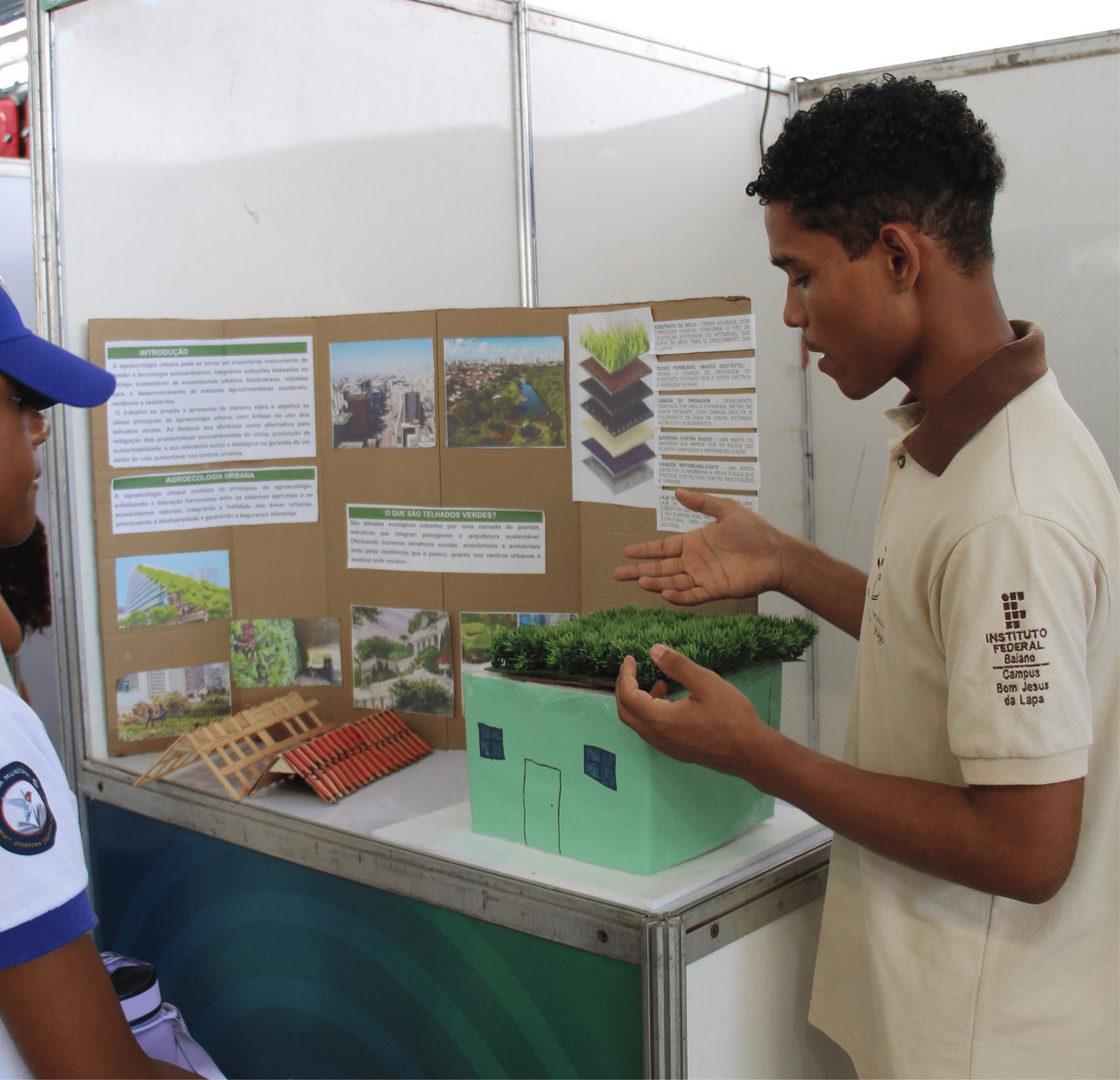 Um jovem negro de cabelo curto e crespo, vestindo uma camisa polo bege com o logo do "Instituto Federal Baiano", demonstra uma maquete. A maquete representa uma casa verde com um "telhado verde" feito de grama sintética. Ao fundo, há um painel de papelão com fotos de cidades, diagramas de camadas de solo e textos sobre "Agroecologia Urbana" e "Telhados Verdes". Uma pessoa de boné azul observa à esquerda.