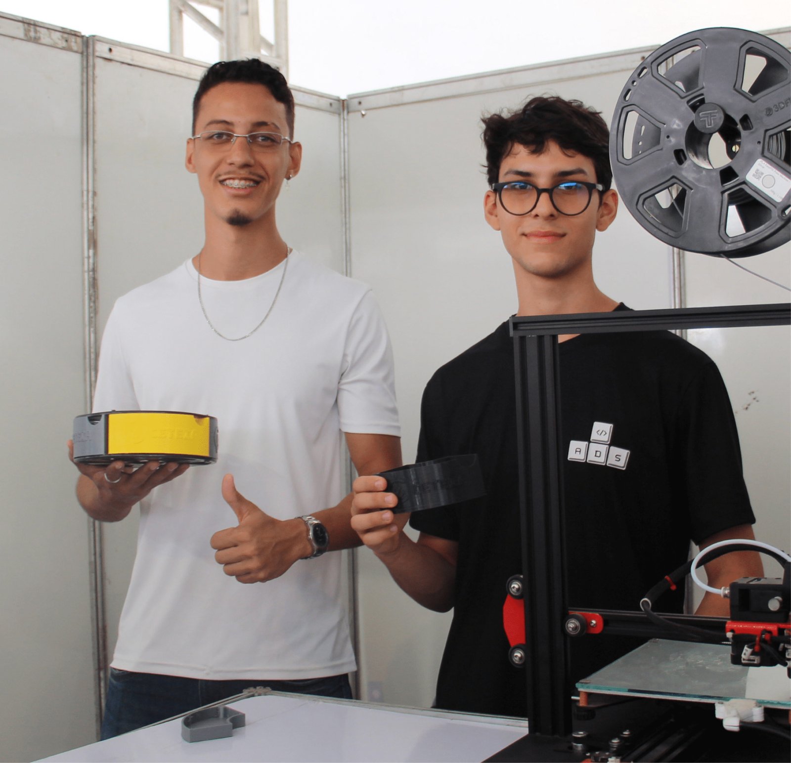 Dois rapazes sorriem atrás de uma impressora 3D em funcionamento. O da esquerda, de camiseta branca, óculos e aparelho, faz um sinal de "joinha". O da direita, de camiseta preta e óculos, segura uma peça preta impressa. No topo da impressora, há um carretel de filamento plástico cinza. A estrutura da impressora é preta com detalhes vermelhos.