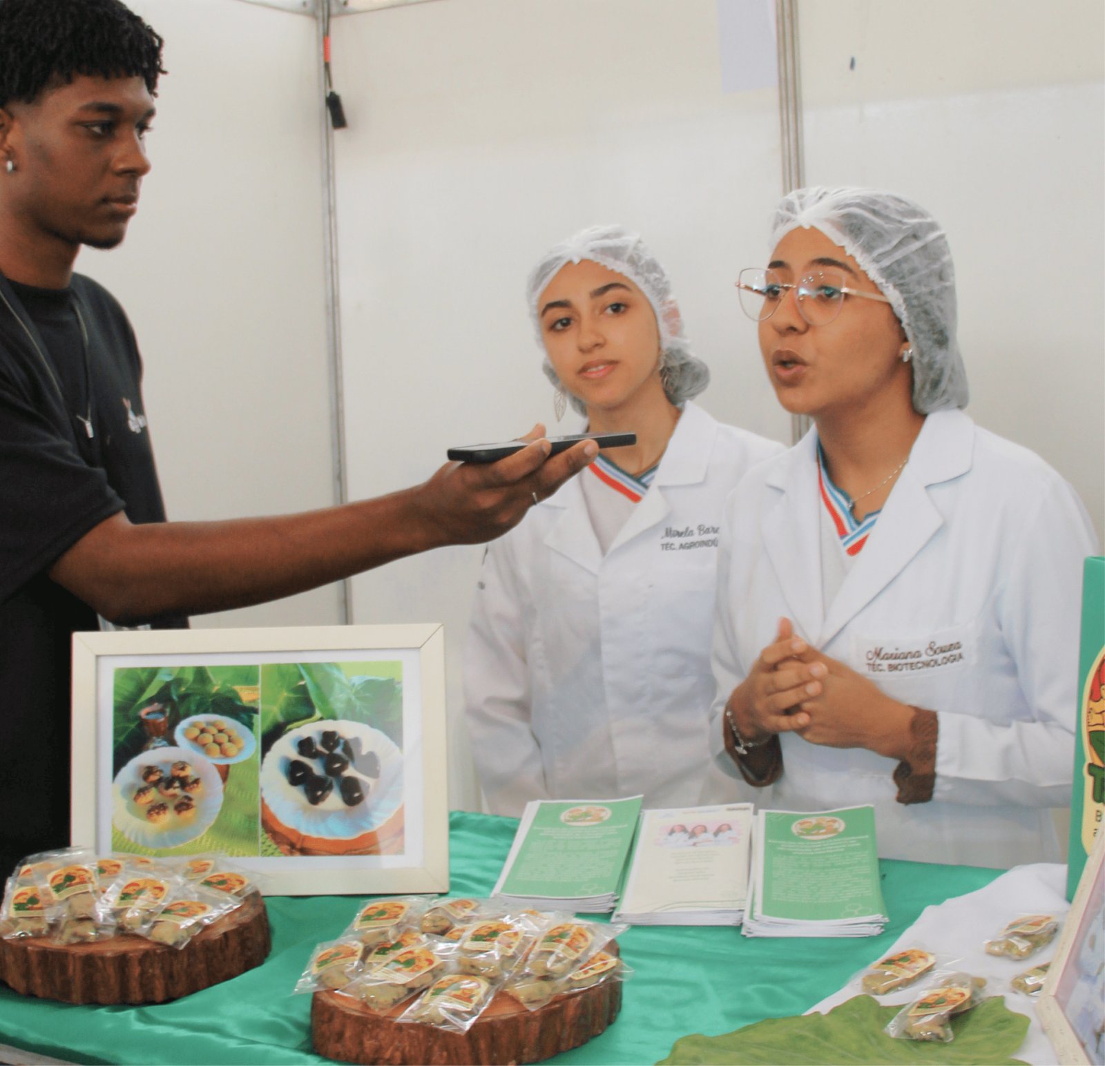Duas jovens estudantes vestindo jalecos brancos, toucas descartáveis e óculos apresentam produtos em um estande. A da direita fala com as mãos gesticulando. À frente delas, sobre uma toalha verde, há discos de madeira servindo como base para diversos produtos embalados (parecem biscoitos ou doces artesanais) e folhetos informativos verdes de "Biotecnologia". Um rapaz à esquerda grava a explicação com o celular.
