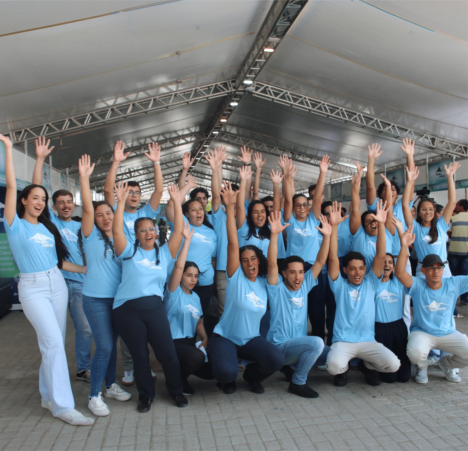 Fotografia em plano aberto sob uma enorme tenda de estrutura metálica. Cerca de 25 jovens, homens e mulheres negros e pardos, vestem camisetas idênticas azul-celeste com a logomarca branca de uma tenda e o texto "Tenda da Ciência". Eles estão eufóricos: muitos têm os braços erguidos e mãos abertas, sorrisos largos e expressões de celebração. Uma jovem à esquerda veste calça branca, enquanto a maioria usa jeans escuros. O chão é de blocos de concreto cinza.