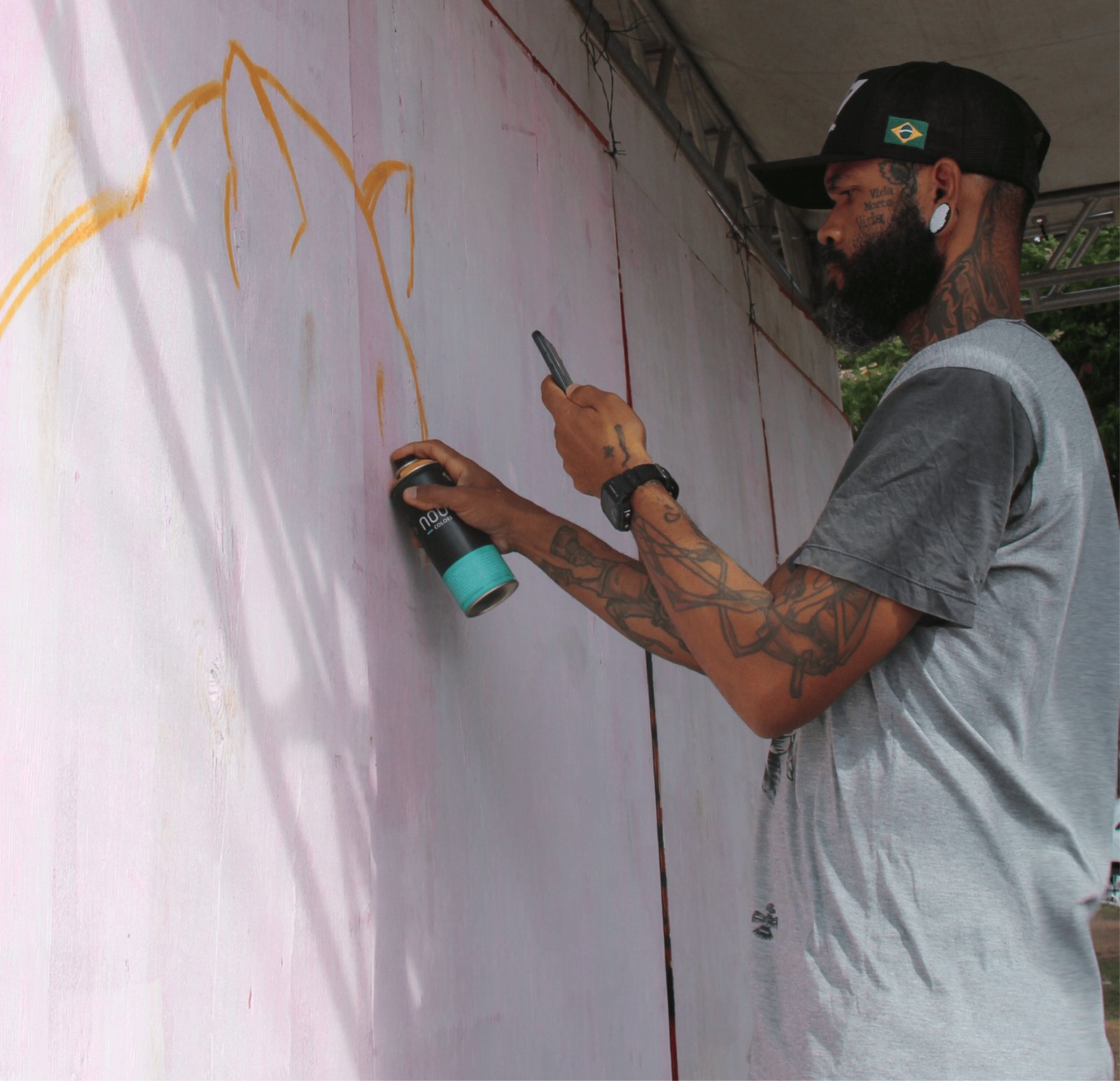 Um homem de perfil, com barba fechada, boné preto com a bandeira do Brasil e braços totalmente tatuados, está grafitando um painel. Ele segura uma lata de spray preta e turquesa da marca "NOU Colors" e usa a outra mão para olhar referências no celular. No painel branco, ele traçou linhas finas em spray amarelo que formam o contorno de uma montanha ou tenda.