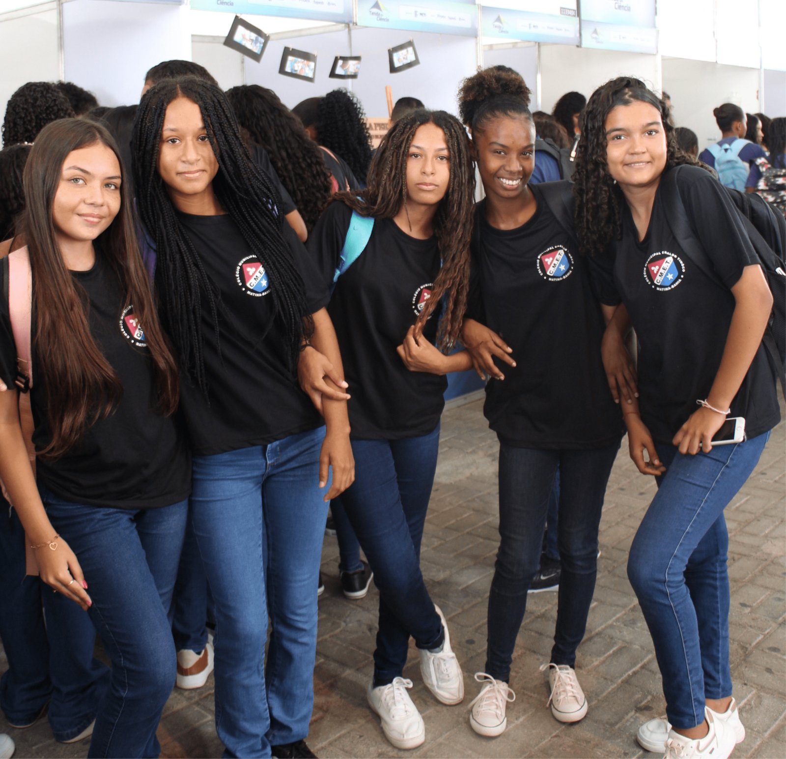Cinco adolescentes negras posam juntas sorrindo. Elas vestem camisetas pretas idênticas com o escudo do "Colégio Municipal Eraldo Tinoco - Matina-Bahia" e calças jeans azuis. Suas peles variam em tons de retinto a pardo, e os cabelos mostram diferentes estilos: tranças nagô longas, cacheados livres e lisos. Elas estão em um corredor de estandes brancos.