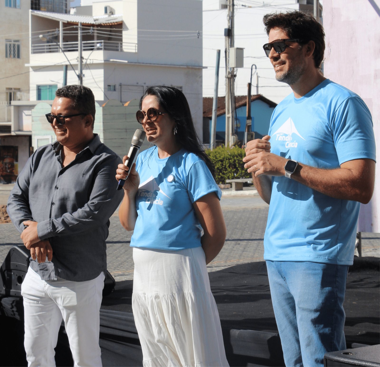 rês adultos estão em um palco ao ar livre. Ao centro, uma mulher de cabelos longos e óculos de sol redondos fala ao microfone; ela usa camiseta azul "Tenda da Ciência" e saia branca longa. À sua esquerda, um homem de camisa cinza e óculos escuros sorri. À direita, um homem alto de óculos escuros, relógio inteligente e camiseta azul observa a fala. Ao fundo, prédios brancos e fiação elétrica sob um céu azul.