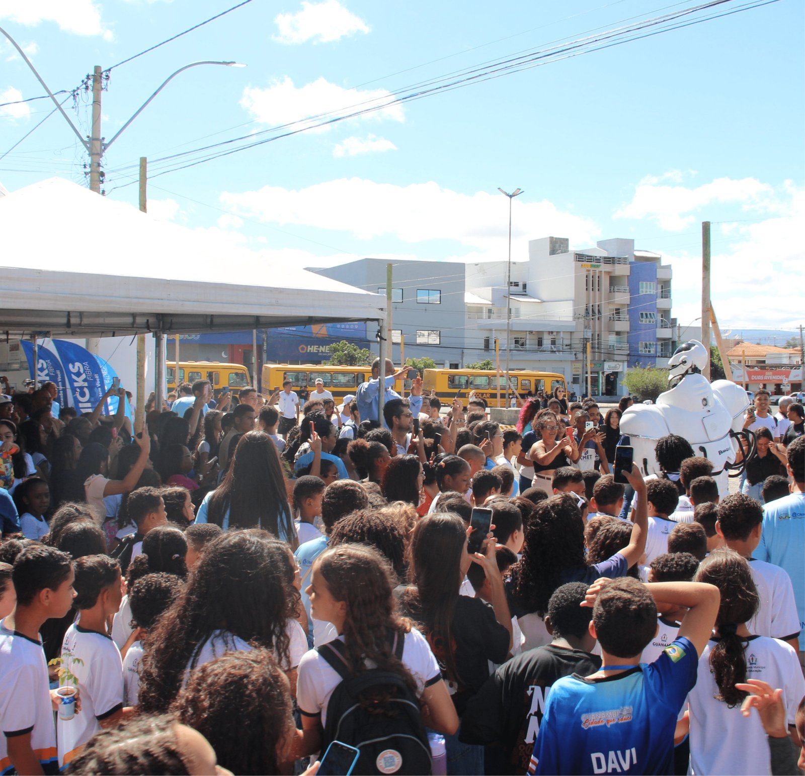 Uma visão panorâmica de uma praça lotada de estudantes (centenas deles). Muitos seguram celulares para o alto, filmando. No lado direito, o "Robozão" branco se destaca acima das cabeças. Ao fundo, veem-se dois ônibus escolares amarelos estacionados perto de prédios comerciais. O céu está azul com algumas nuvens brancas, e o sol brilha forte.