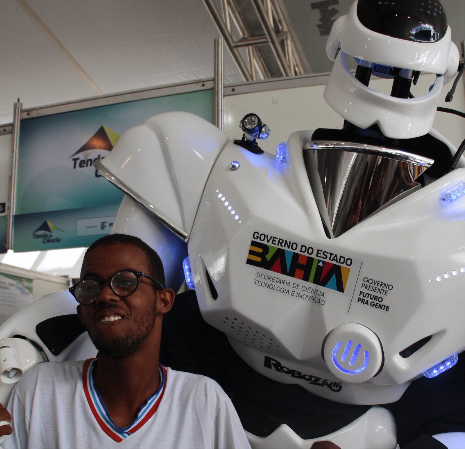 Um jovem negro de óculos e barba rala sorri radiante para a foto. Ele está posicionado logo à frente do grande Robozão branco, que parece "abraçá-lo" com seu braço mecânico gigante. O jovem usa uma camiseta branca com detalhes em azul e vermelho na gola. A imagem captura um momento de pura alegria e inclusão tecnológica.