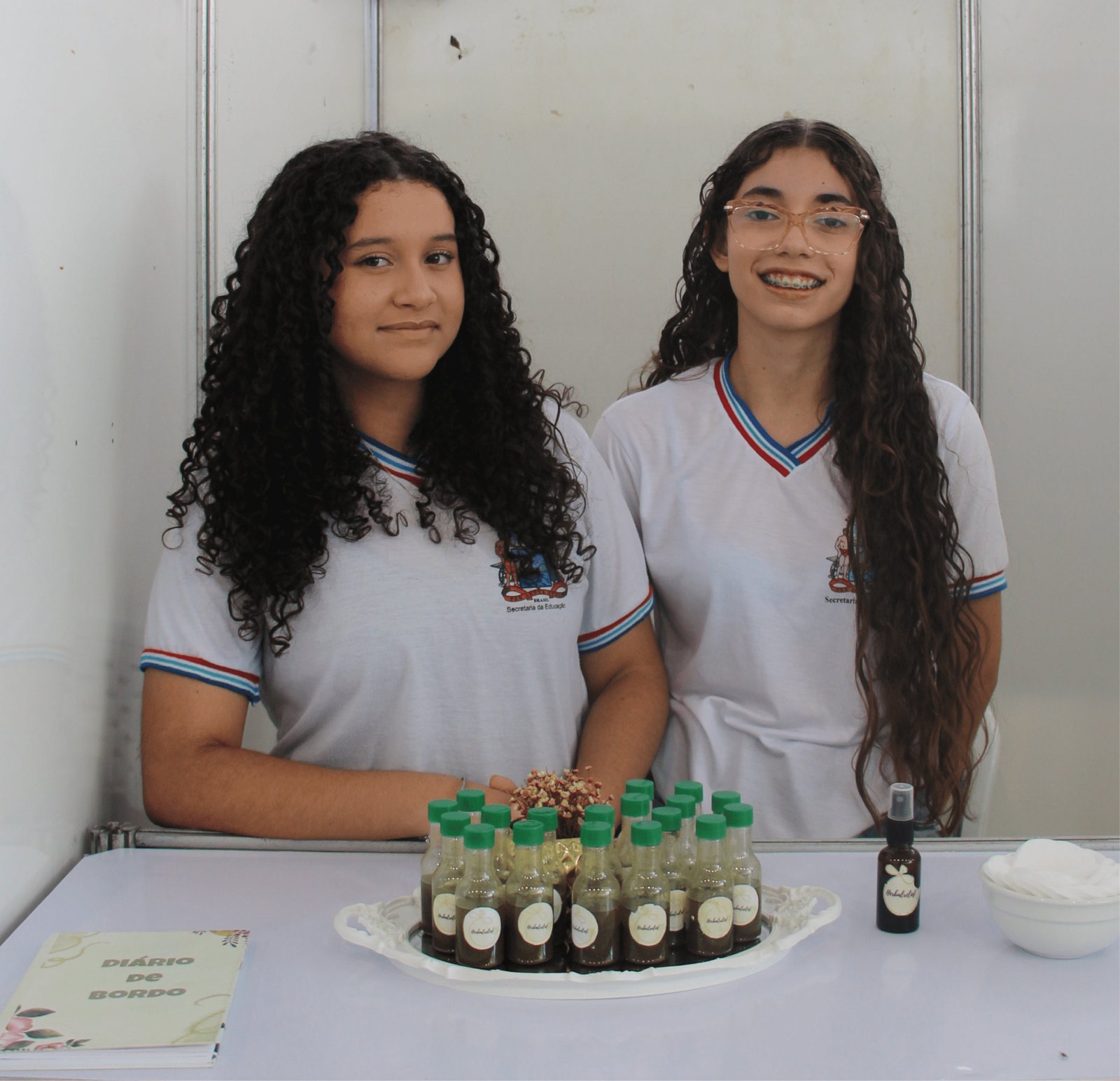 Duas estudantes de cabelos longos e cacheados, usando uniformes brancos com gola em V listrada (azul e vermelho), posam atrás de uma mesa. Elas apresentam uma bandeja branca com dezenas de pequenos frascos de vidro contendo um líquido escuro (provavelmente extratos vegetais) com rótulos circulares. Há também um frasco de spray maior e um caderninho intitulado "DIÁRIO DE BORDO" com ilustrações florais.