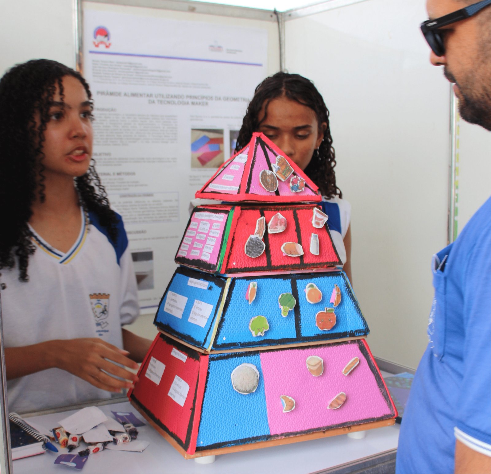Duas jovens demonstram uma "Pirâmide Alimentar Maker". A pirâmide é uma estrutura 3D grande feita de placas coloridas (rosa, azul e verde) com texturas de borracha. Em cada nível, há ícones recortados de alimentos: pães e arroz na base, frutas e vegetais no meio, carnes e leite acima, e doces no topo. Um homem de óculos e camisa azul observa a explicação atentamente à direita.